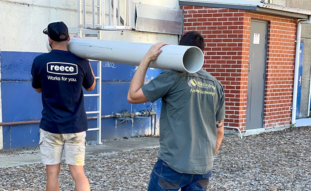 Two pipe relining employees carrying a big pipe on their shoulders